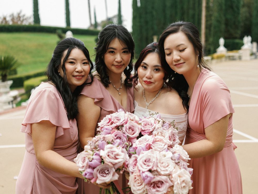 Bridesmaids in pink dresses huddle together while holding pink and white floral bouquets during outdoor wedding photos.