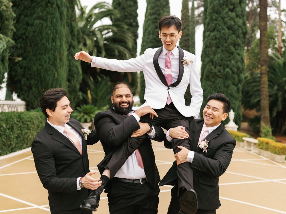 A group of groomsmen in black suits playfully pose and jump during a wedding photo session in a garden setting.