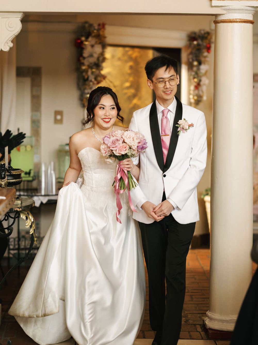 Couple in white wedding attire walking through a bright hallway with classical columns and decor.