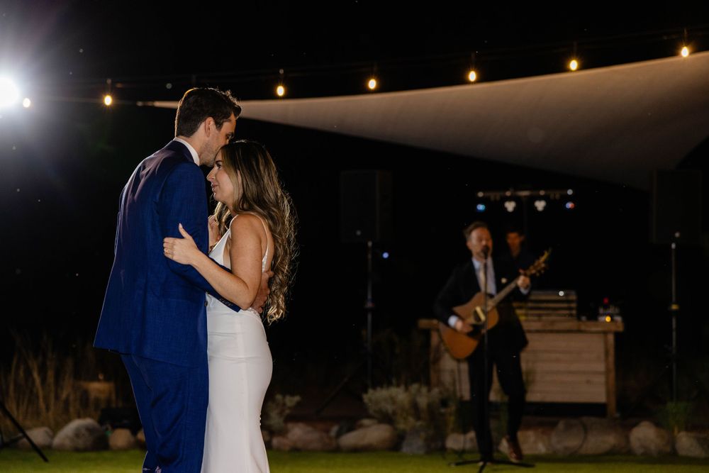 Couple shares first dance at outdoor reception while musician performs in background.
