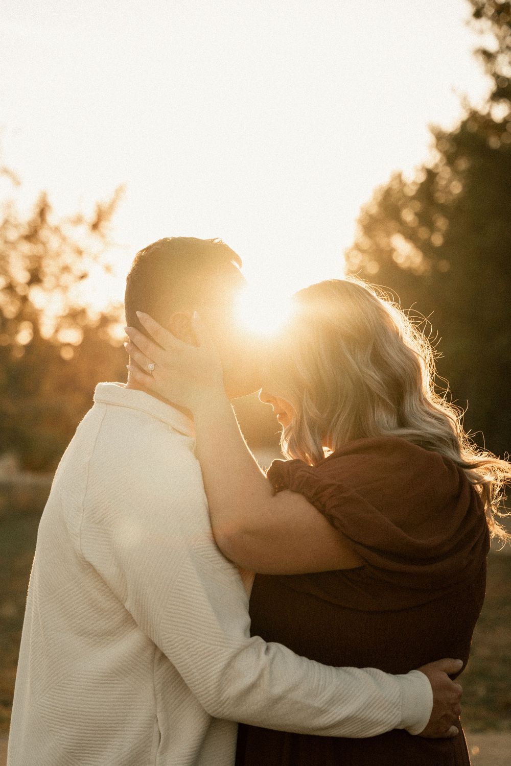 A couple shares a romantic embrace during golden hour sunset with sunlight creating a warm backlit glow.