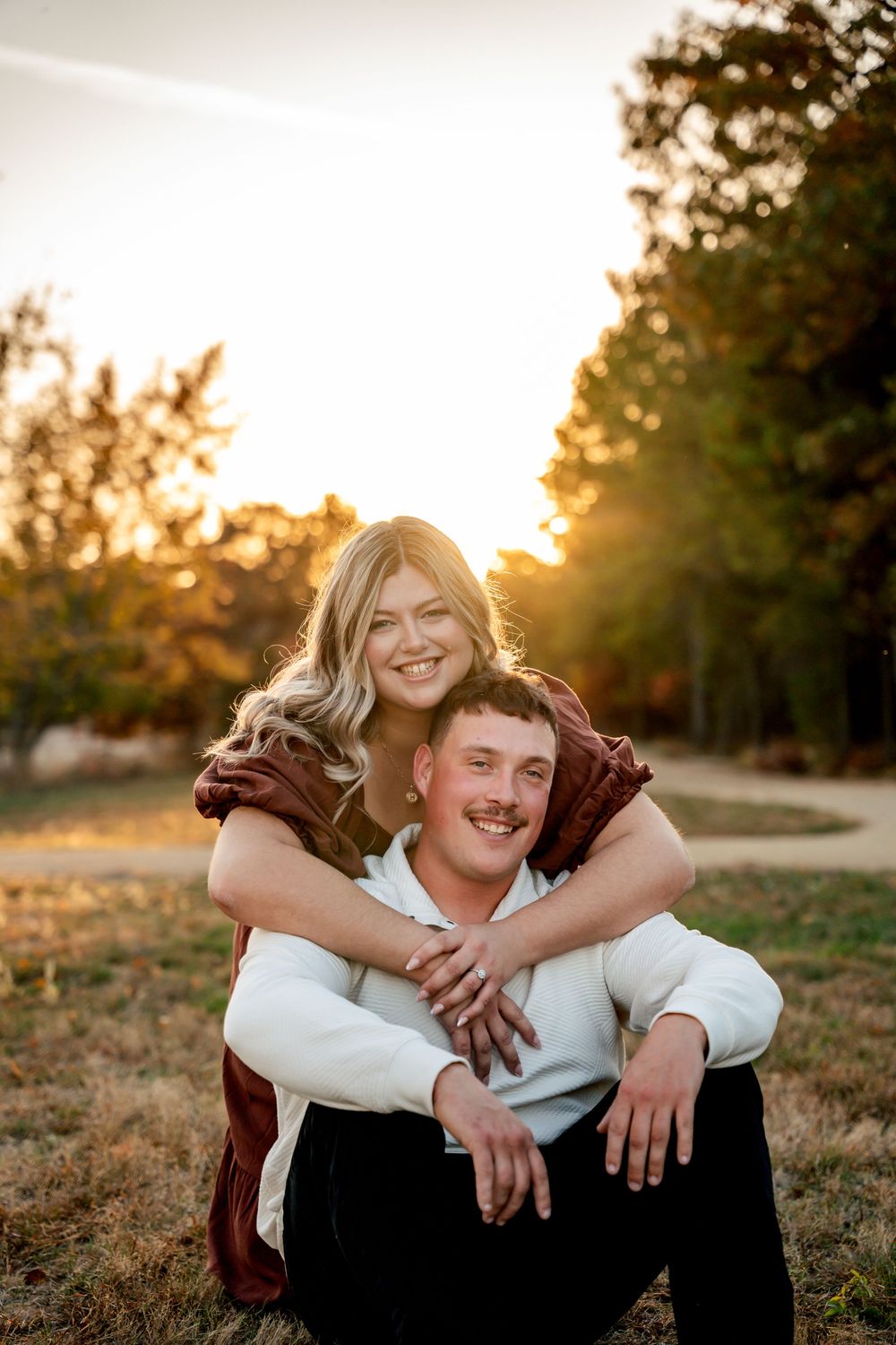 A couple shares a joyful embrace while sitting outdoors during golden hour sunset in a natural setting.