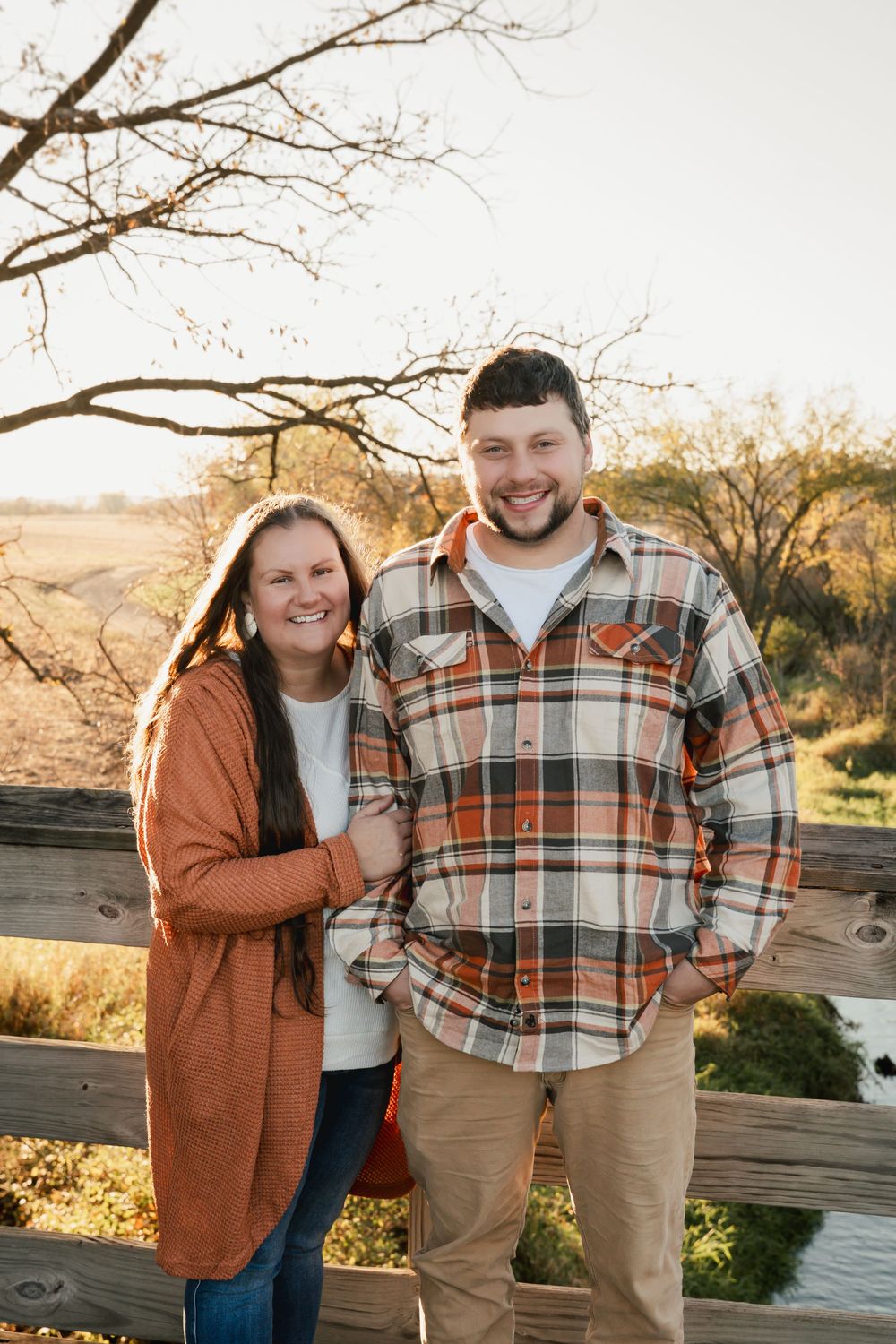 A couple stands together by a wooden fence during golden hour in autumn, wearing casual layers and plaid.