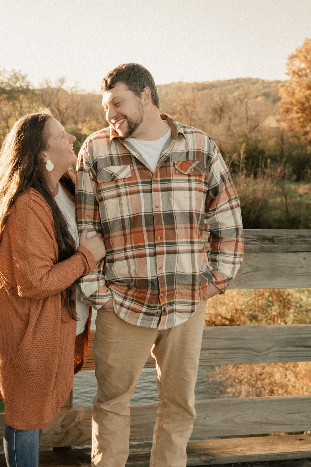 A couple in autumn attire shares playful moments outdoors during golden hour sunset in a rustic setting.