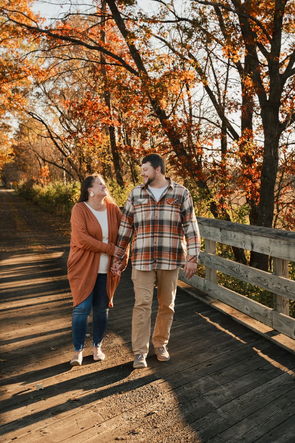 Couple walking along wooden boardwalk surrounded by autumn trees with golden foliage at sunset.
