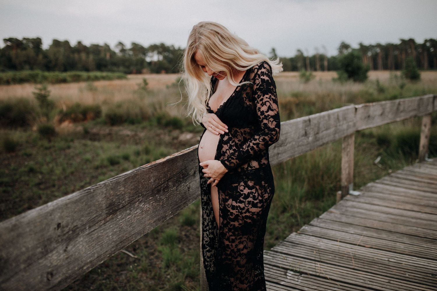 Pregnant woman in black lace gown cradling baby bump on wooden bridge in nature.