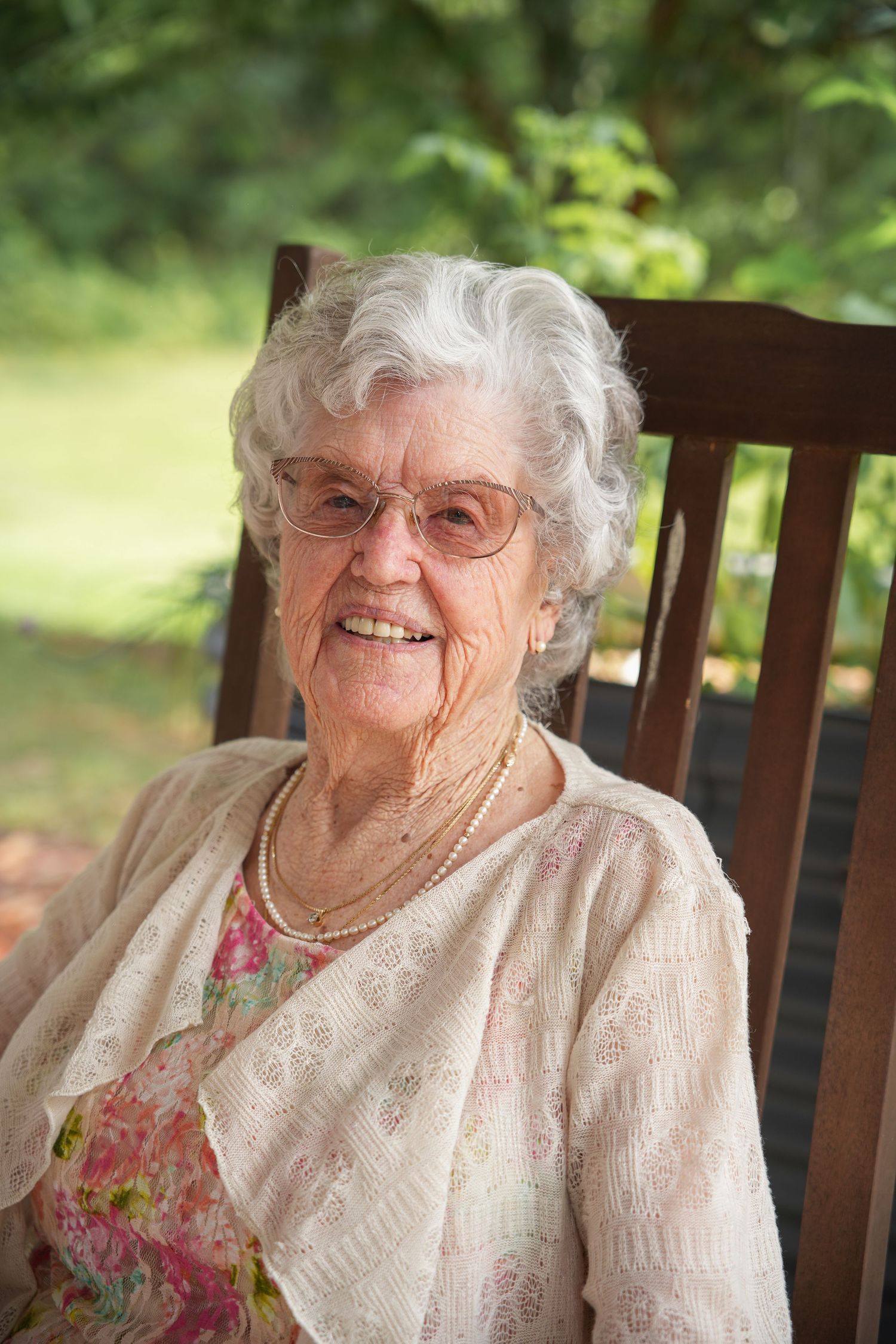 An elderly person in a lace blouse smiles warmly while sitting on a wooden bench outdoors.