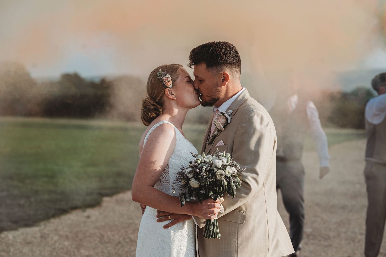 A couple shares a romantic kiss during their outdoor wedding ceremony at sunset while holding an elegant floral bouquet.