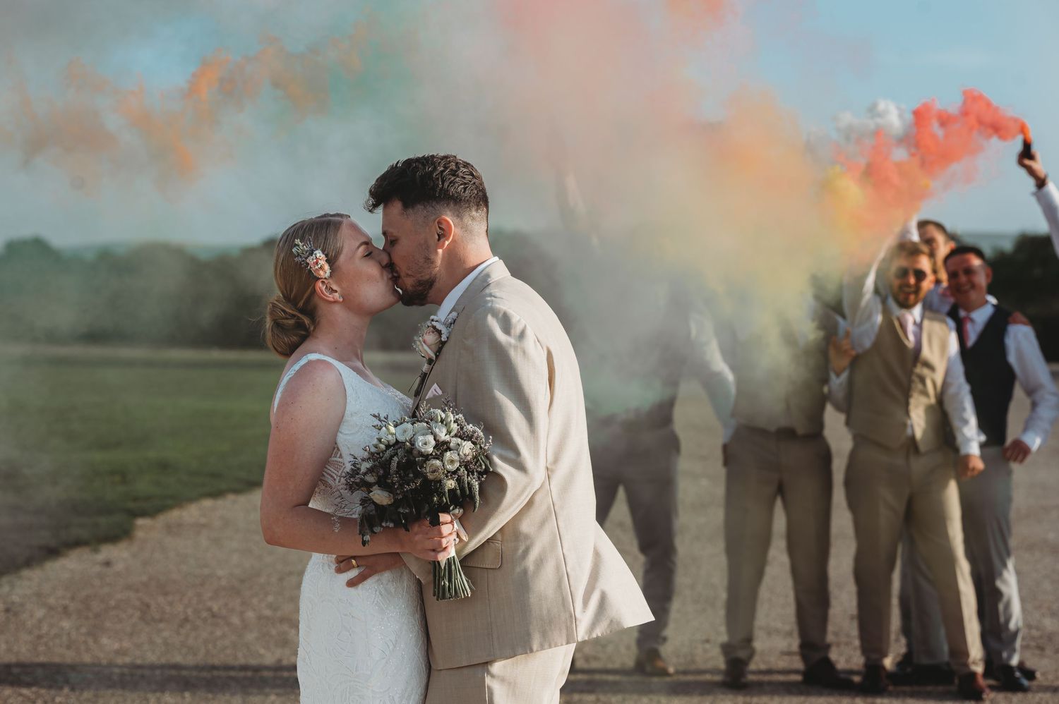 A romantic wedding couple embraces on a countryside path while guests hold colorful smoke flares in the background.