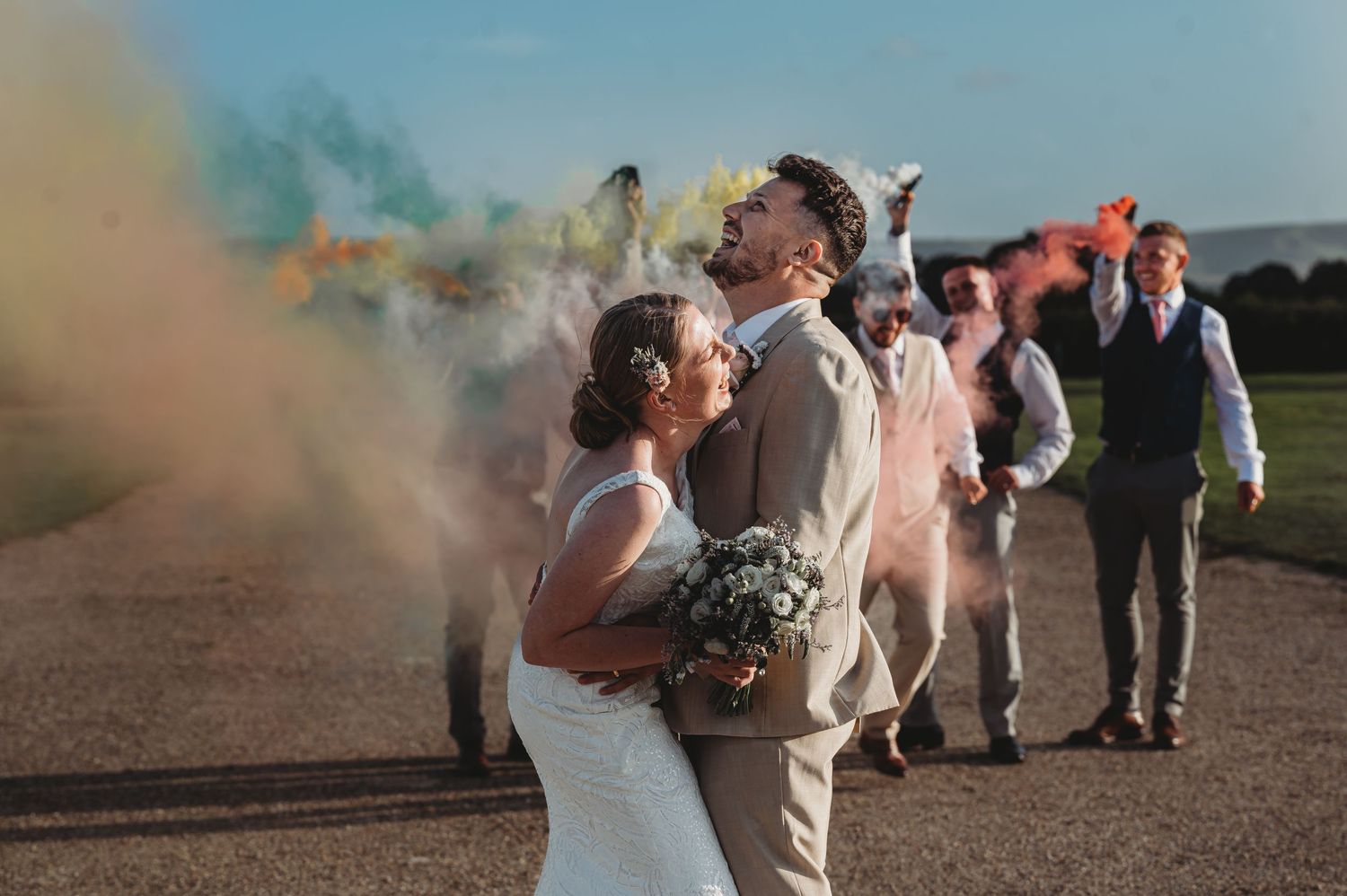 A wedding couple celebrates with smoke bombs creating colorful plumes on a dirt road while guests look on in the background.