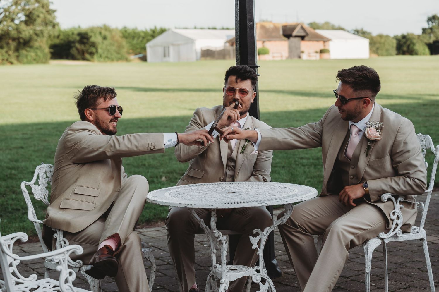 Groomsmen in beige suits celebrating together at an outdoor table with ornate white chairs on a grassy lawn.