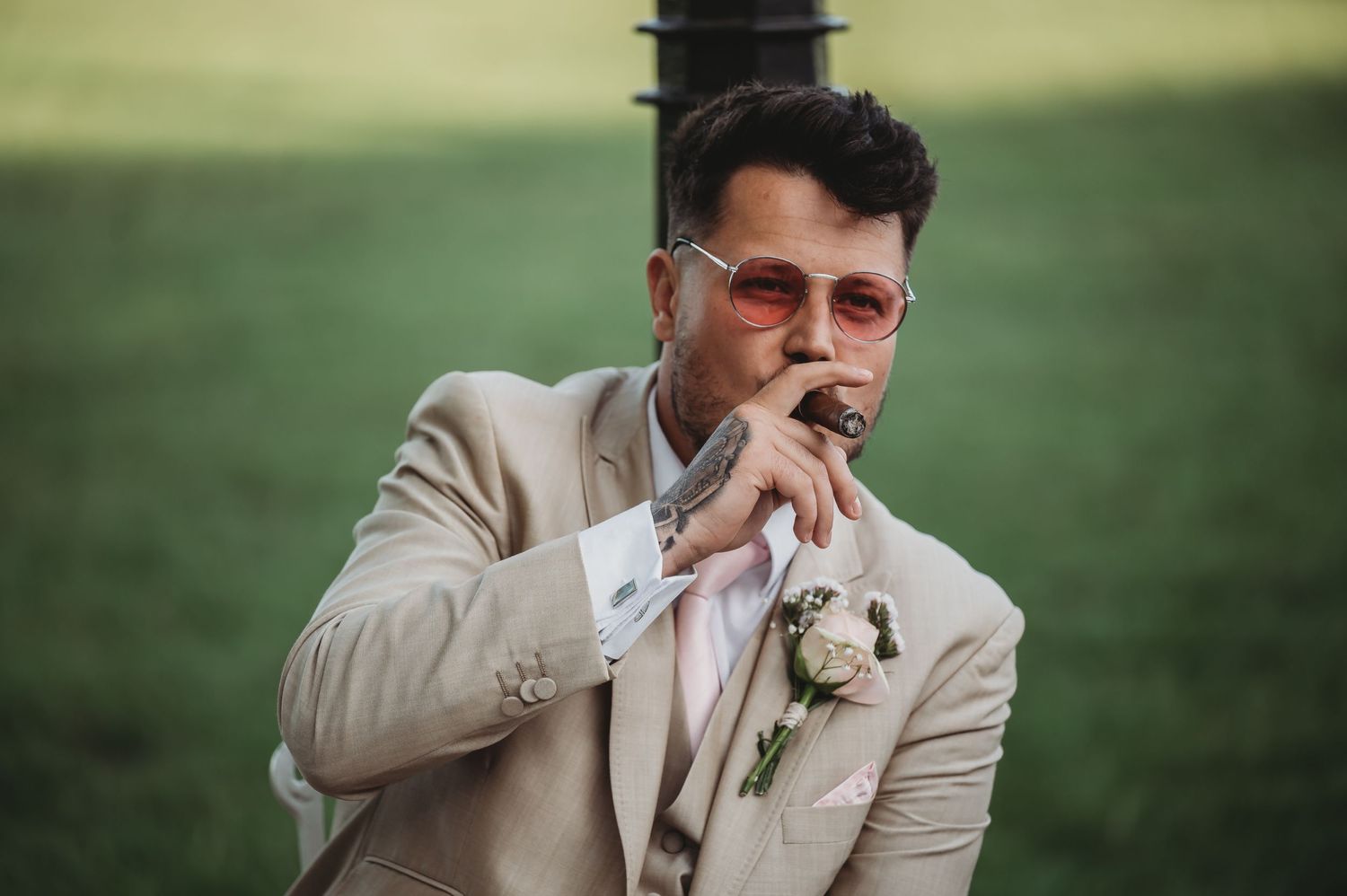Groom in beige suit and sunglasses with boutonniere smokes while posing in outdoor setting at sunset.