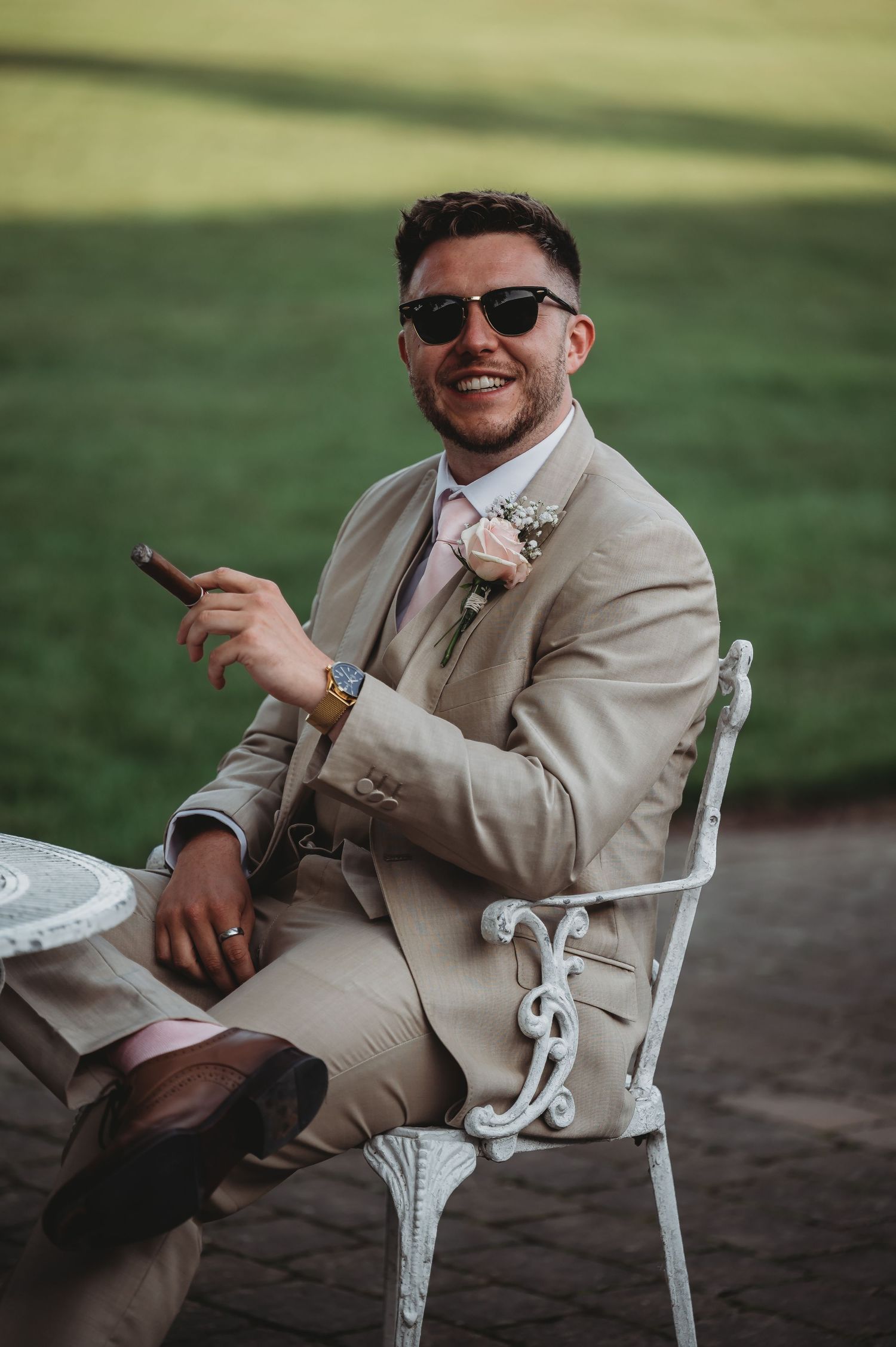 A wedding guest in a beige suit and sunglasses sits in an ornate white garden chair at an outdoor event.