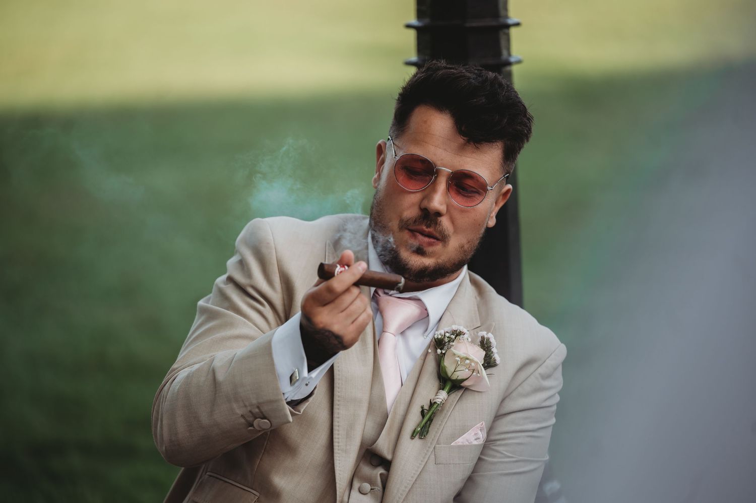 A person in a cream-colored suit jacket and boutonniere points toward the camera against a blurred green background.