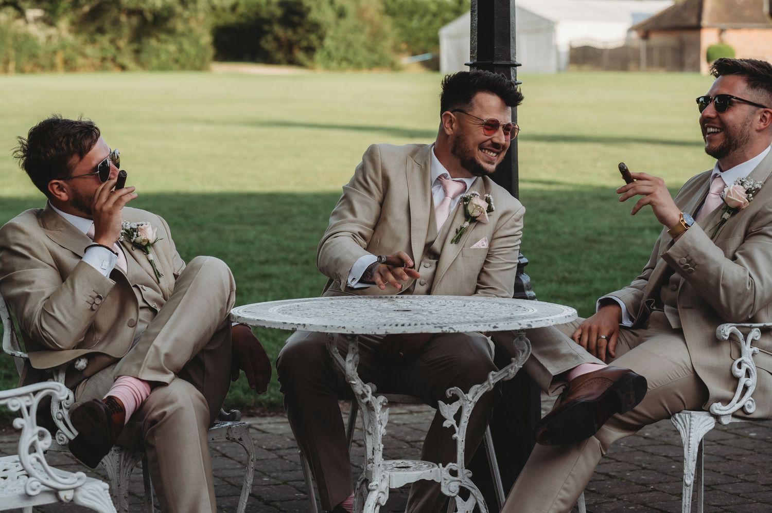 Three people in beige suits share a laugh while seated at an ornate white garden table outdoors.