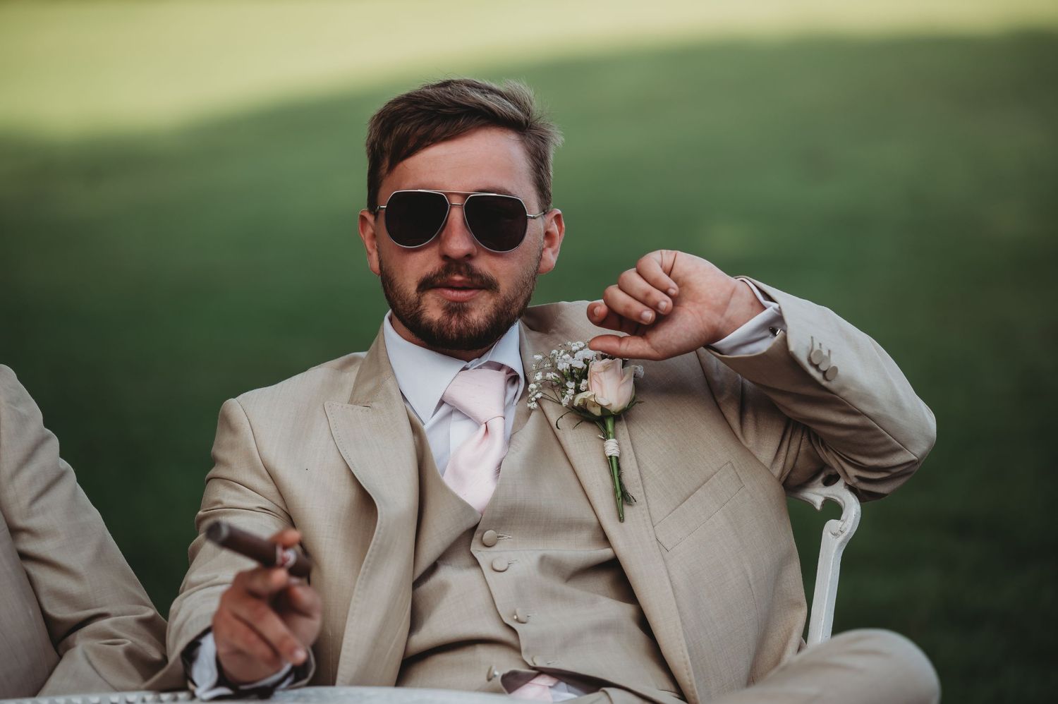 A groomsman in a beige suit and sunglasses relaxes at an outdoor wedding venue.