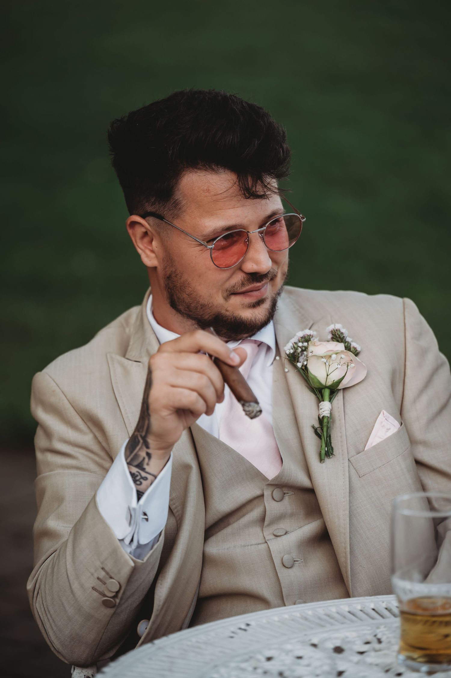 A groom in a beige three-piece suit and rose-tinted glasses holds a cigar while sitting at an outdoor reception.