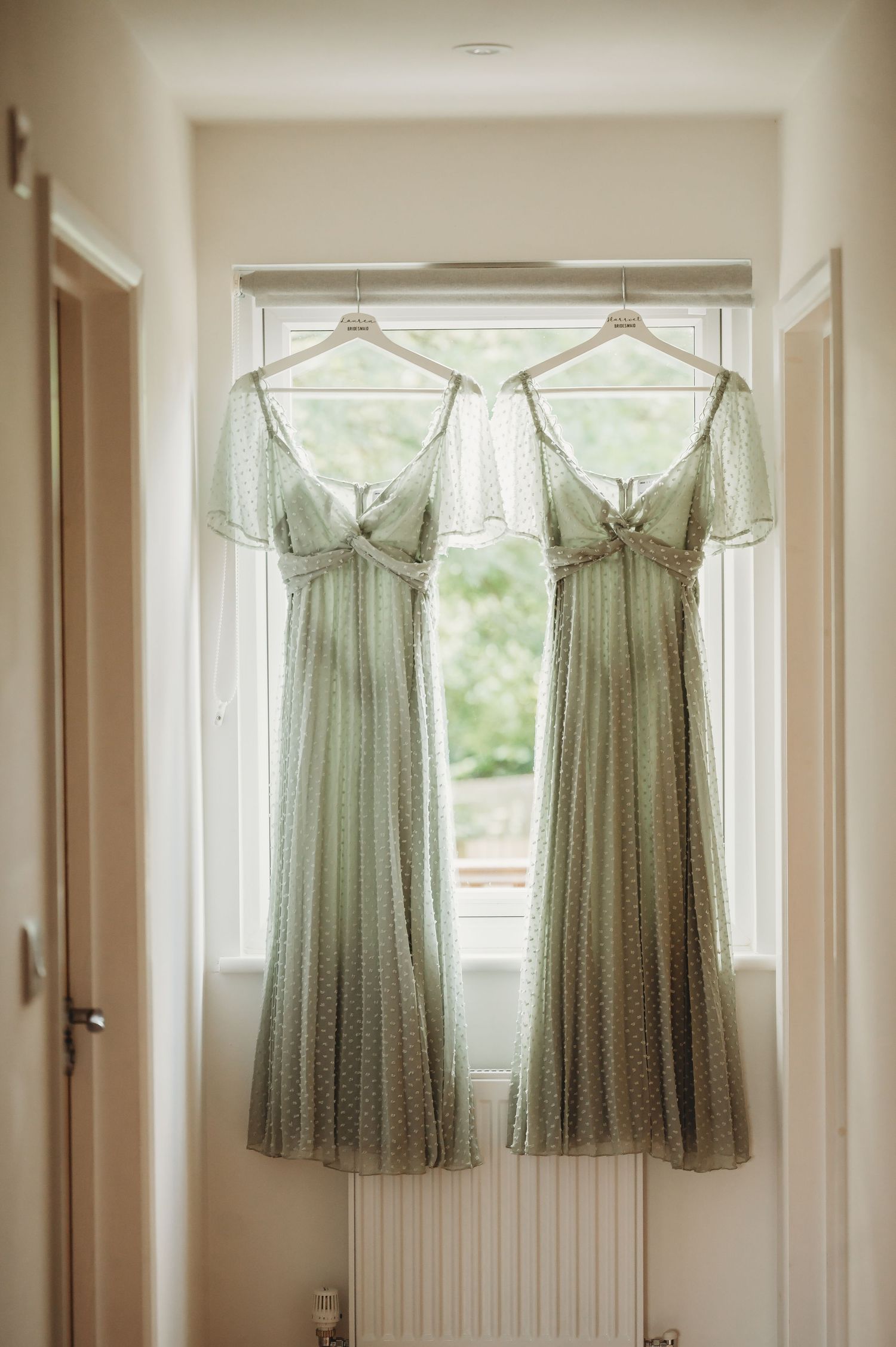 Two sage green bridesmaid dresses with polka dots hanging in a window frame.