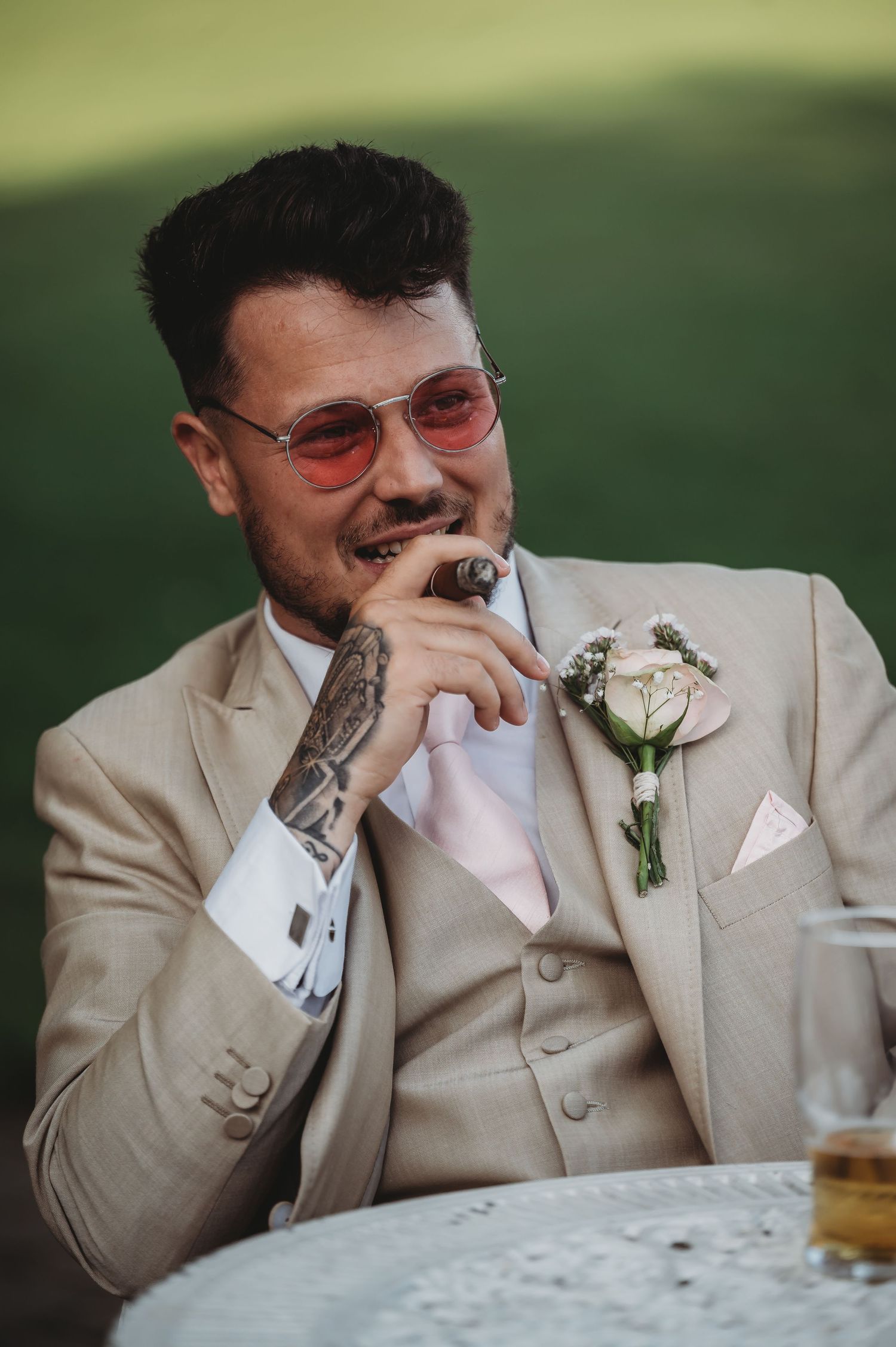 A groom in a beige suit and tinted glasses smiles while wearing a floral boutonniere at a wedding celebration.
