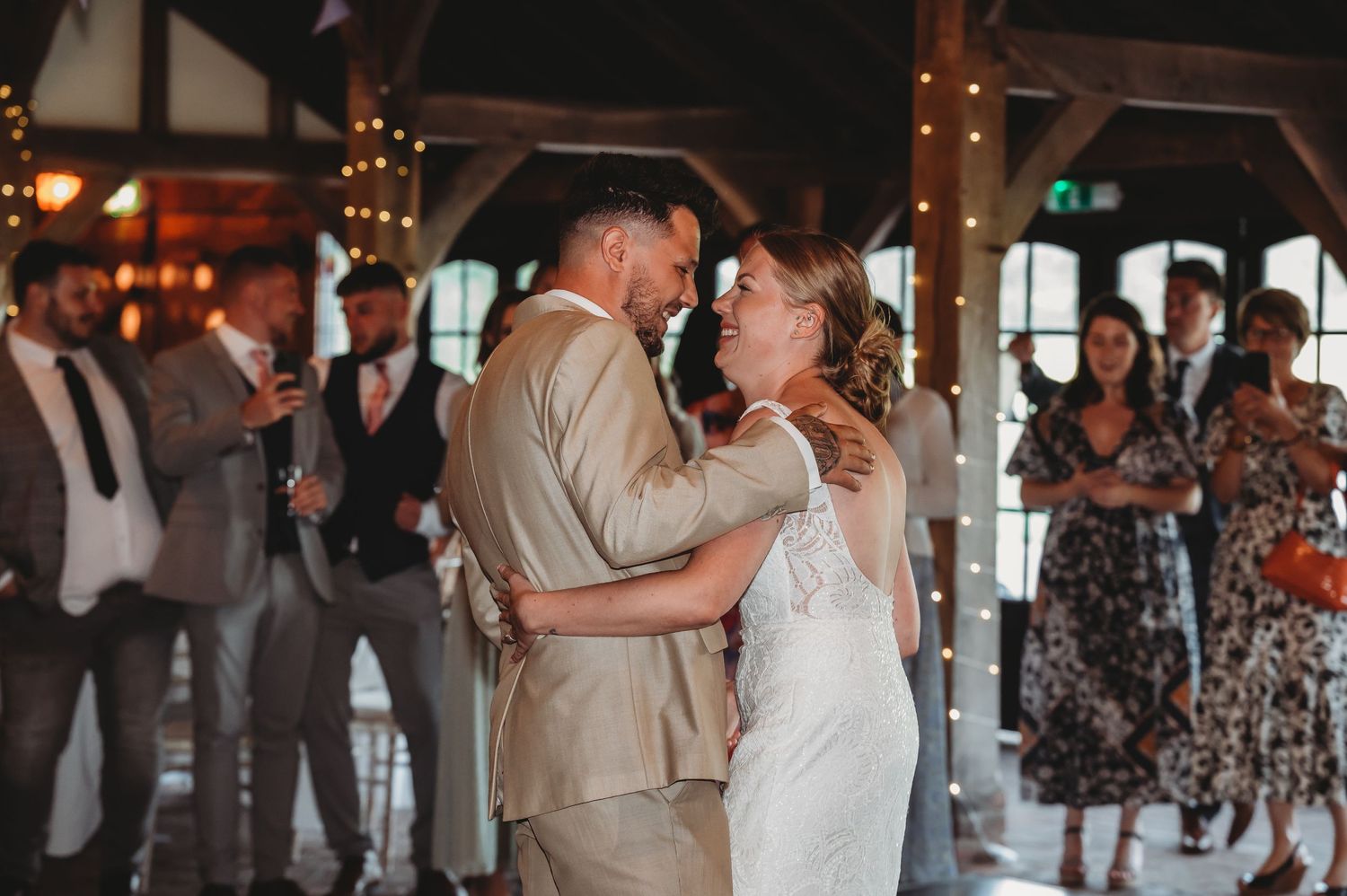 Couple shares their first dance as newlyweds in a beautifully lit rustic venue while guests look on.