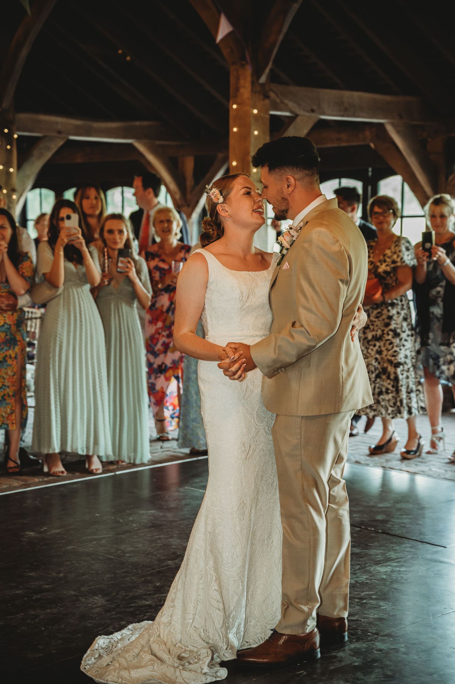 A couple shares their first dance at a rustic wedding venue while guests in pastel dresses look on.