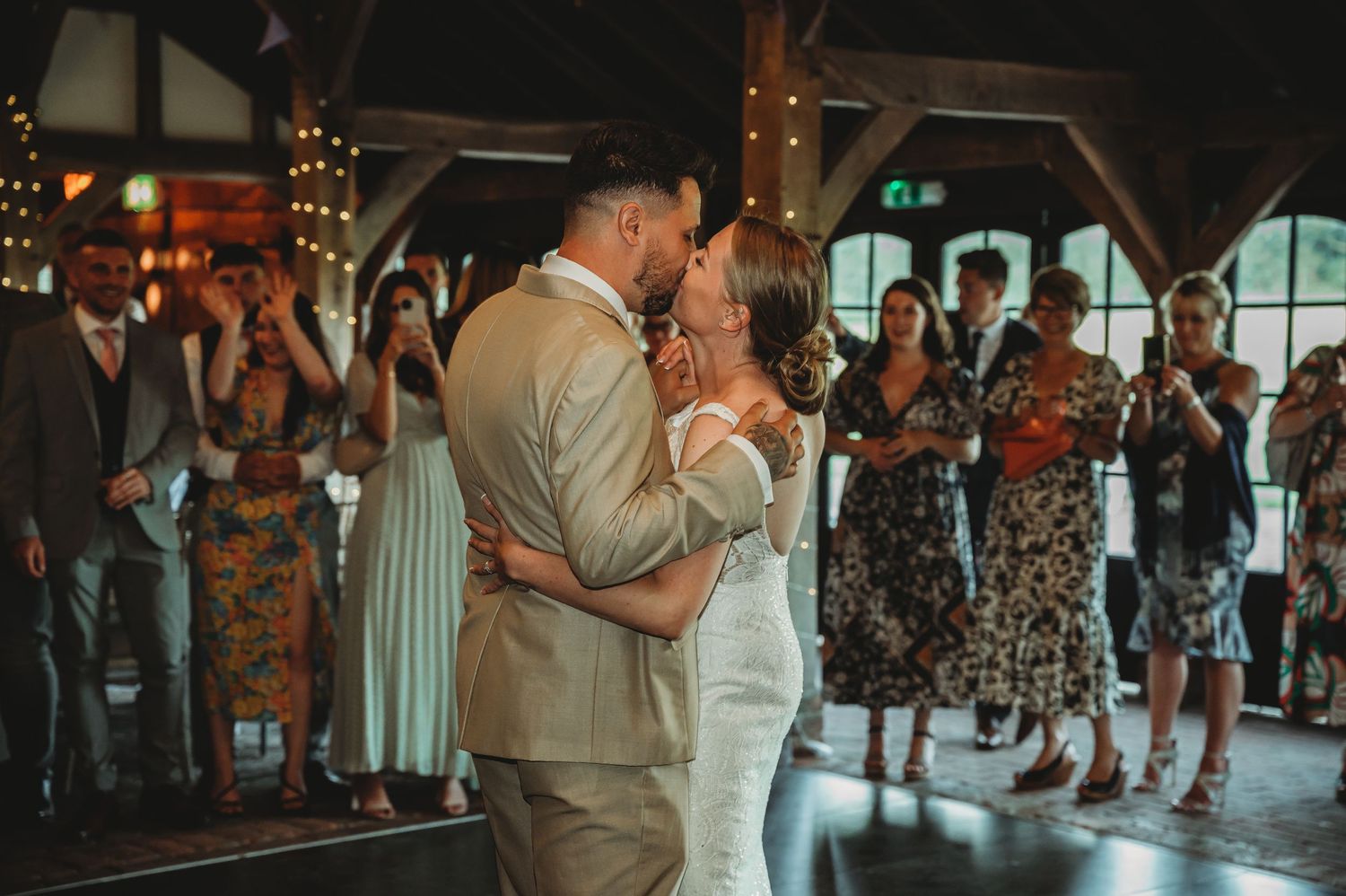 Newlyweds share their first dance surrounded by guests at an indoor reception with string lights and rustic decor.