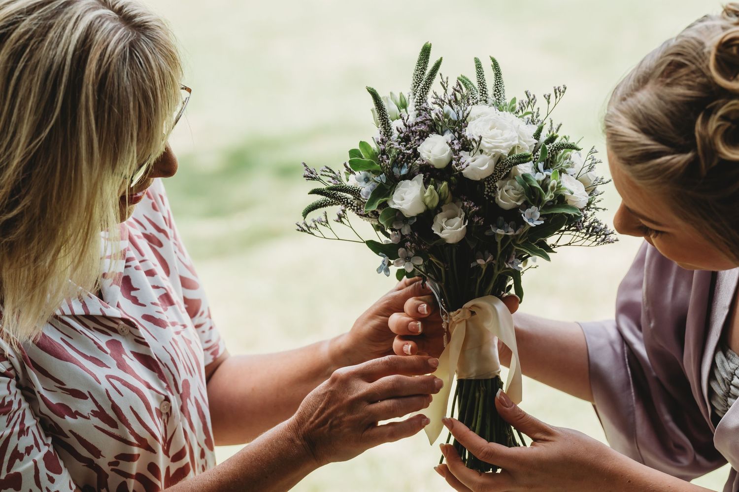 Two people look at a beautiful white and green wedding bouquet bound with twine and ribbon against a blurred green backdrop.