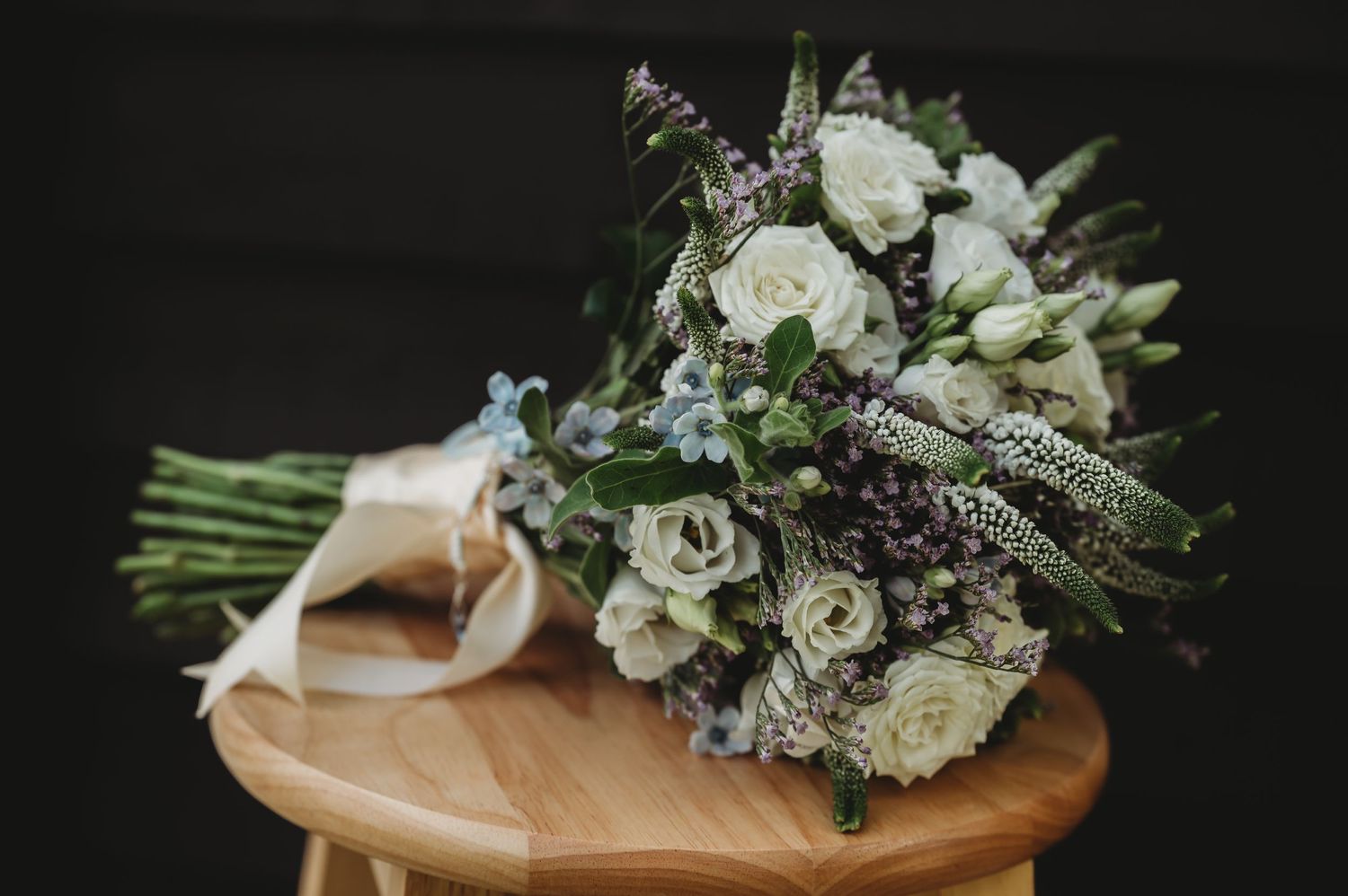 A rustic bridal bouquet with white roses and purple wildflowers tied with ribbon rests on wooden surface.