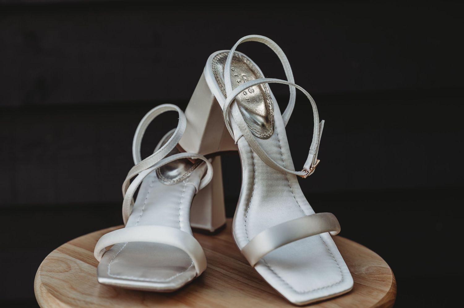 White square-toe bridal sandals with ankle straps displayed on a wooden surface against dark background.