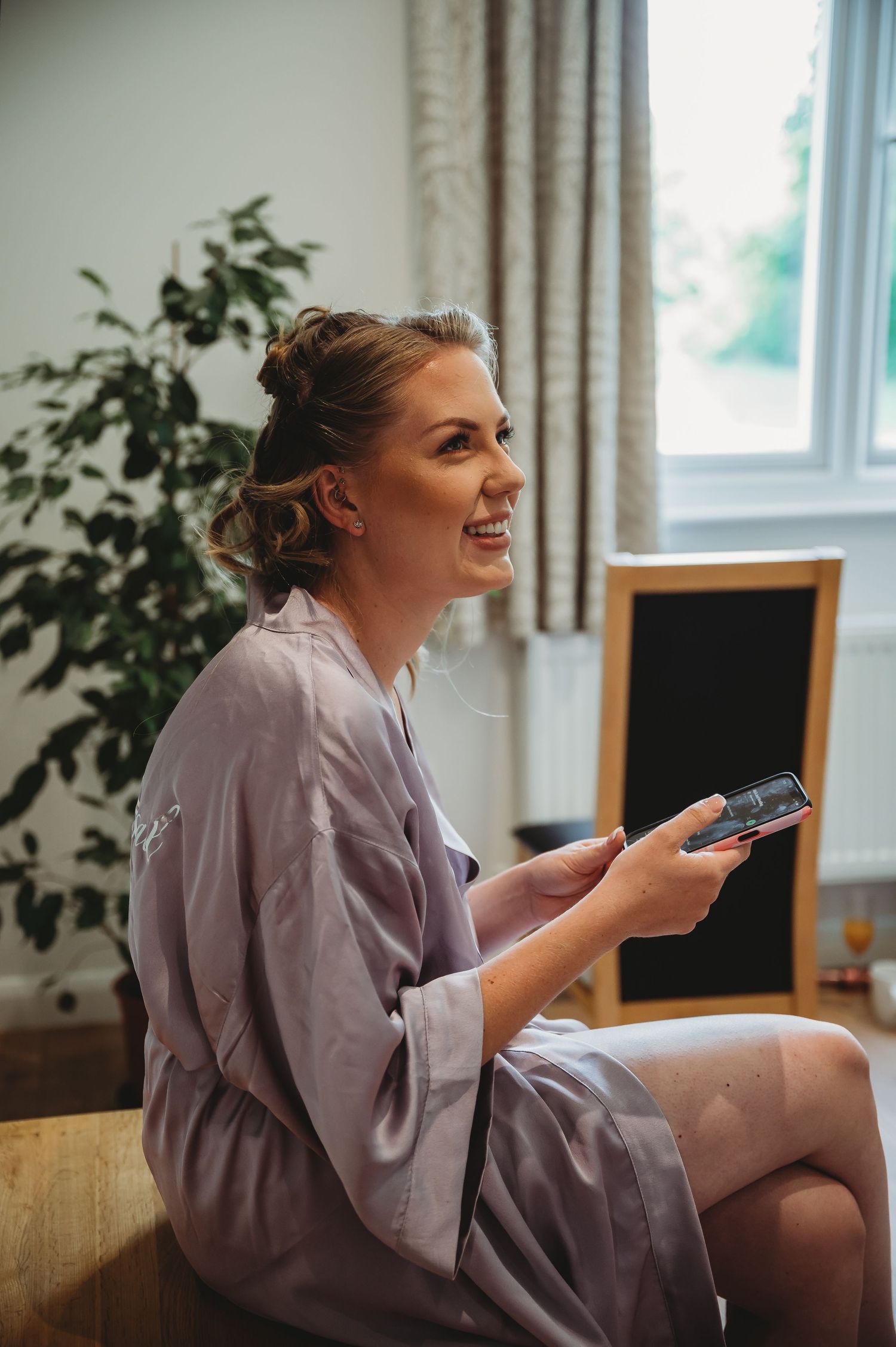 Person in grey robe sits on couch smiling while holding phone in bright living room with houseplants.