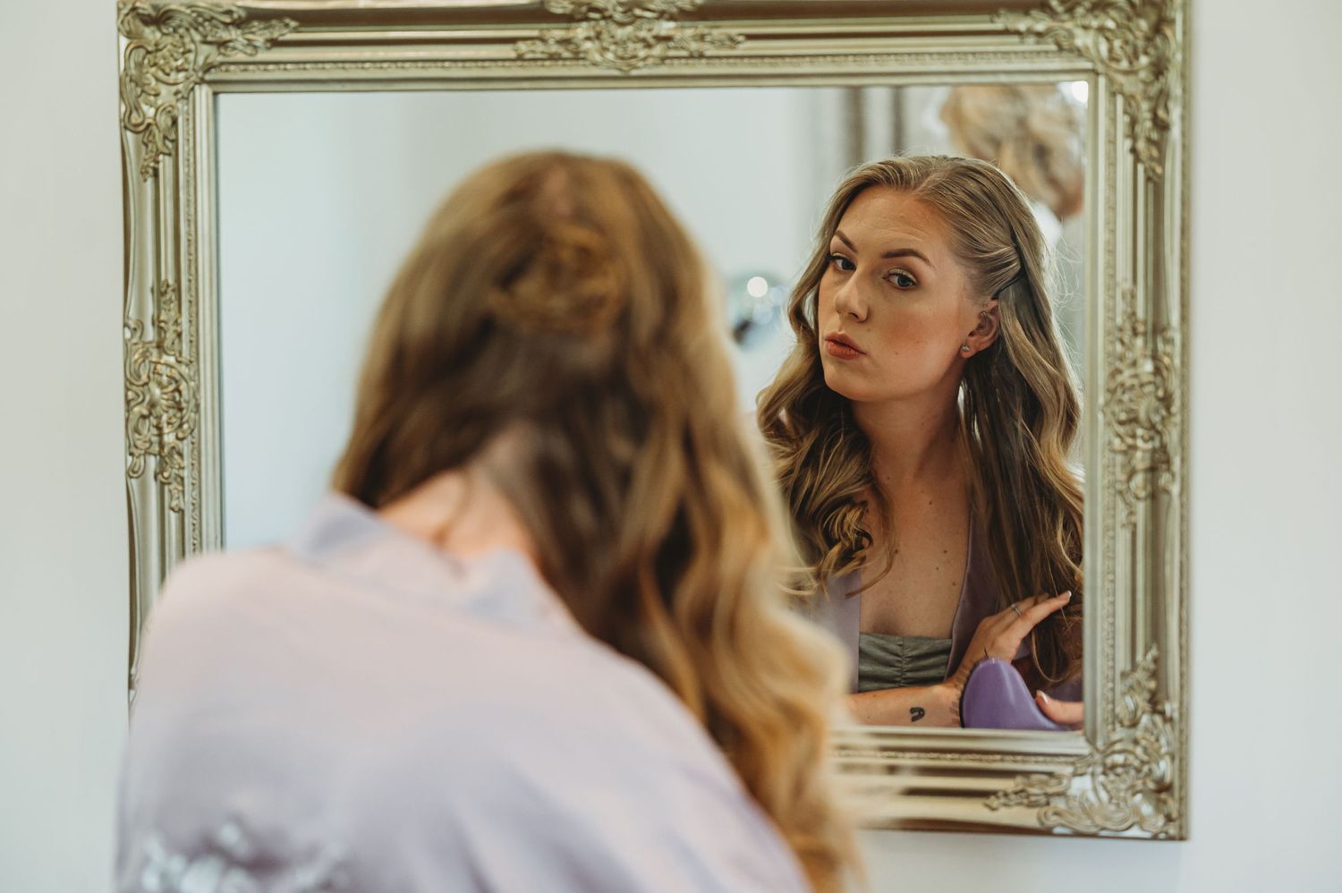 A person gazes at their reflection in an ornate gold-framed mirror mounted on a wall.