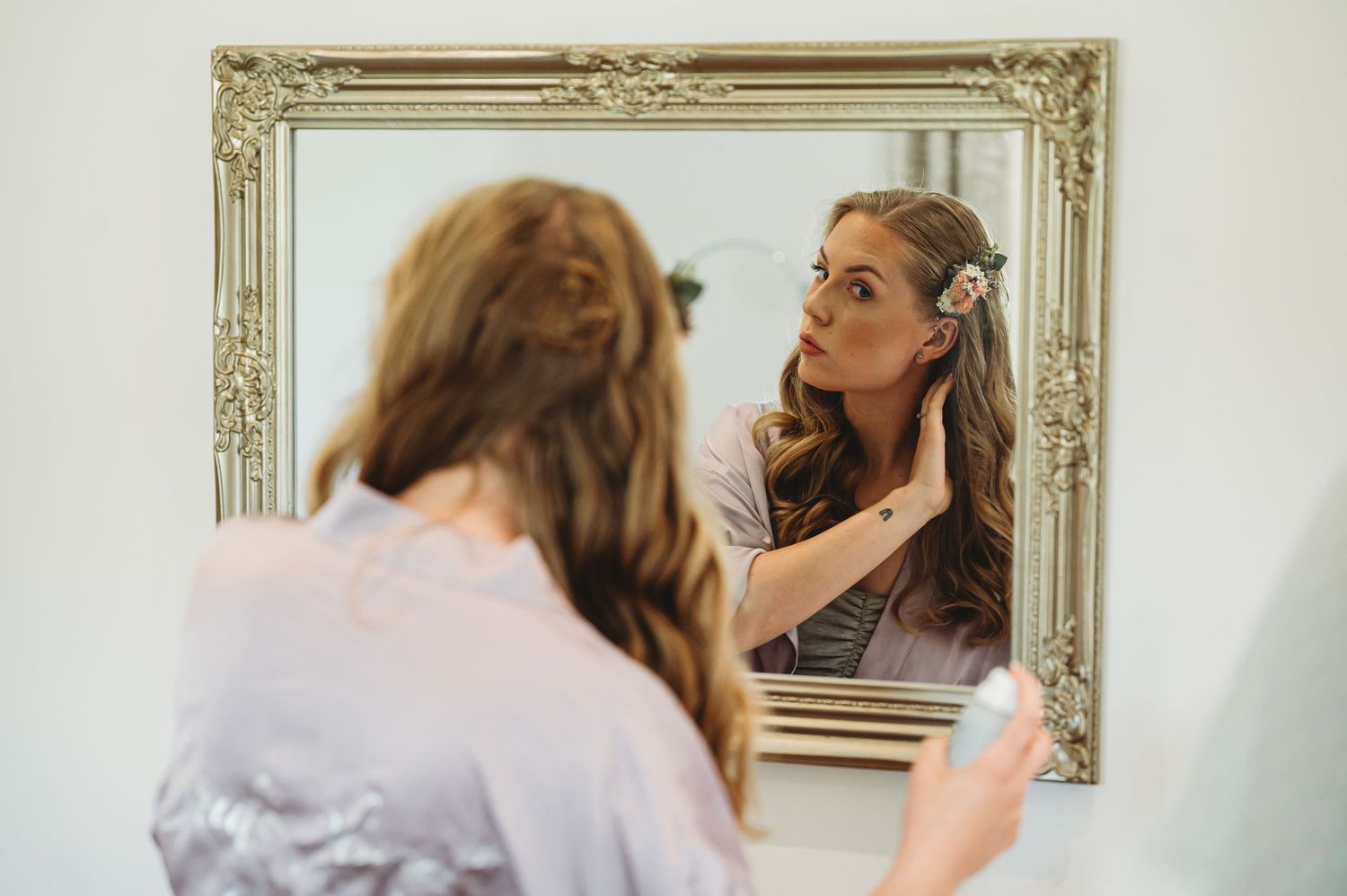 Someone looks in an ornate framed mirror while adjusting their hair in a soft-lit room.