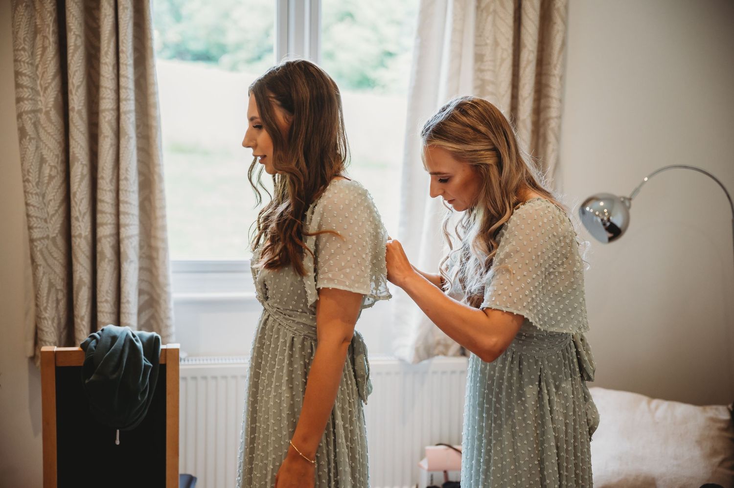 Two women wearing vintage-style gingham dresses prepare for a special occasion in a bright interior room.