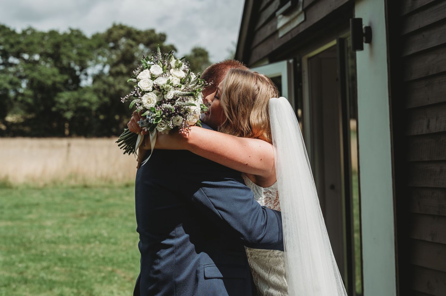 A couple sharing an emotional embrace outdoors during a wedding ceremony with a white bouquet and flowing veil.