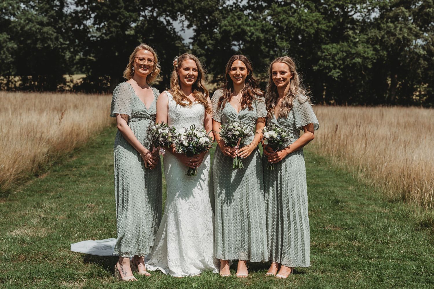 A bridal party wearing soft mint green dresses poses together in a rustic field with tall golden grass and trees.