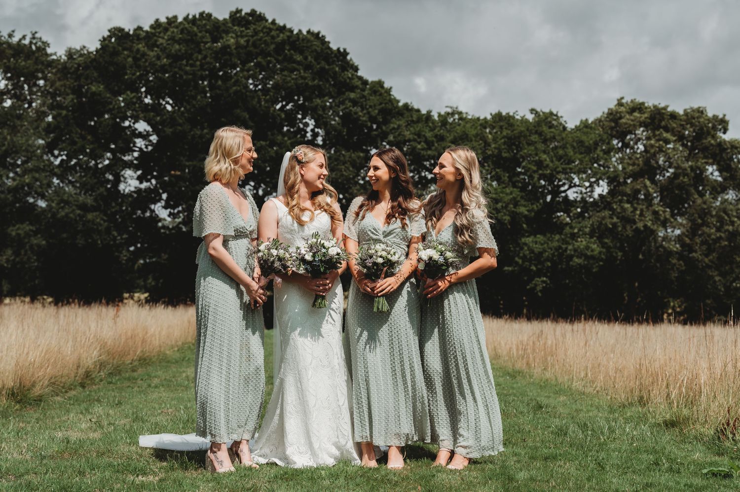 A group in sage green dresses and one in white stand together in a grassy field with trees in the background.