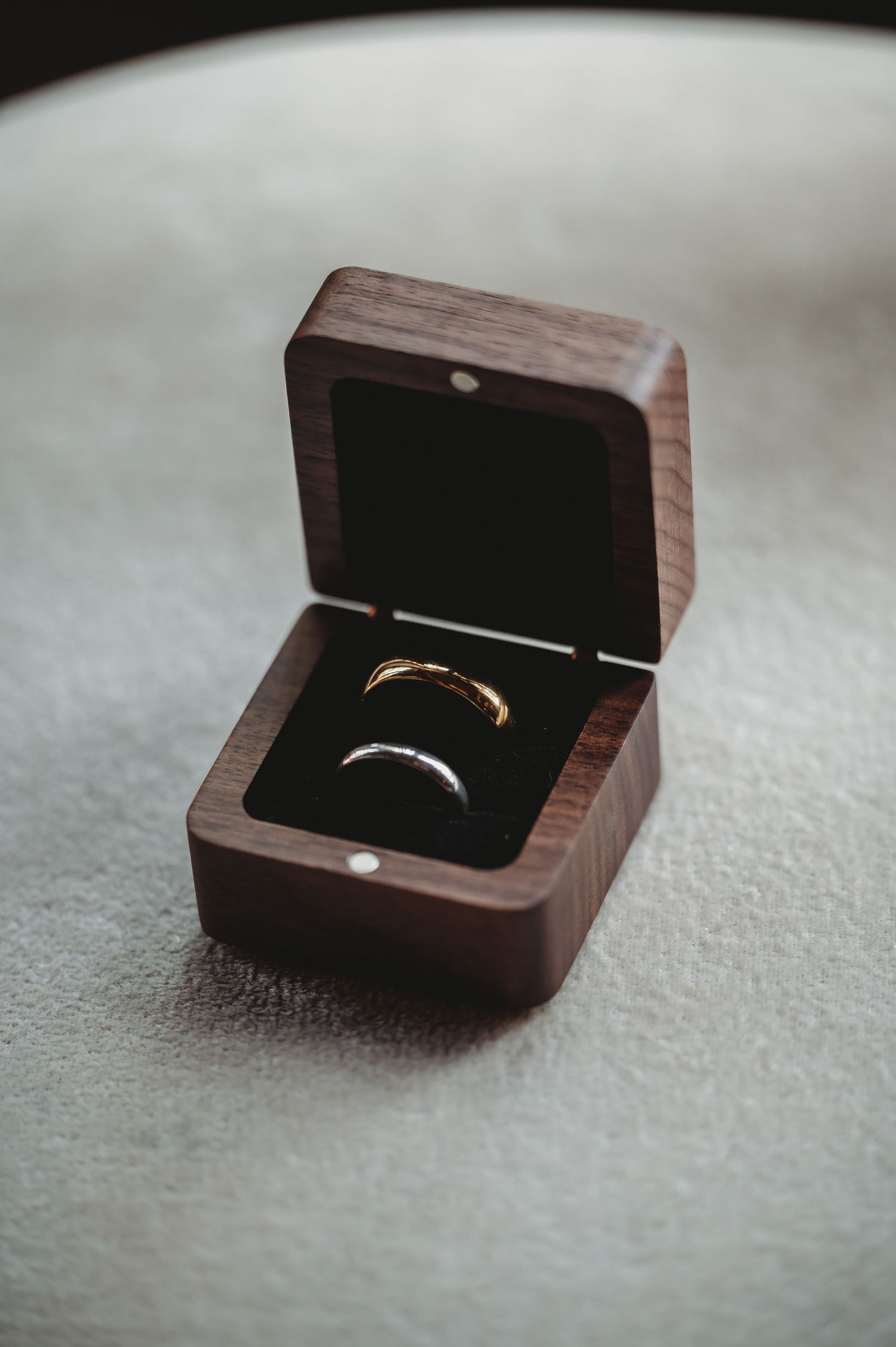 Two wedding rings displayed in an open wooden ring box on a textured gray surface.
