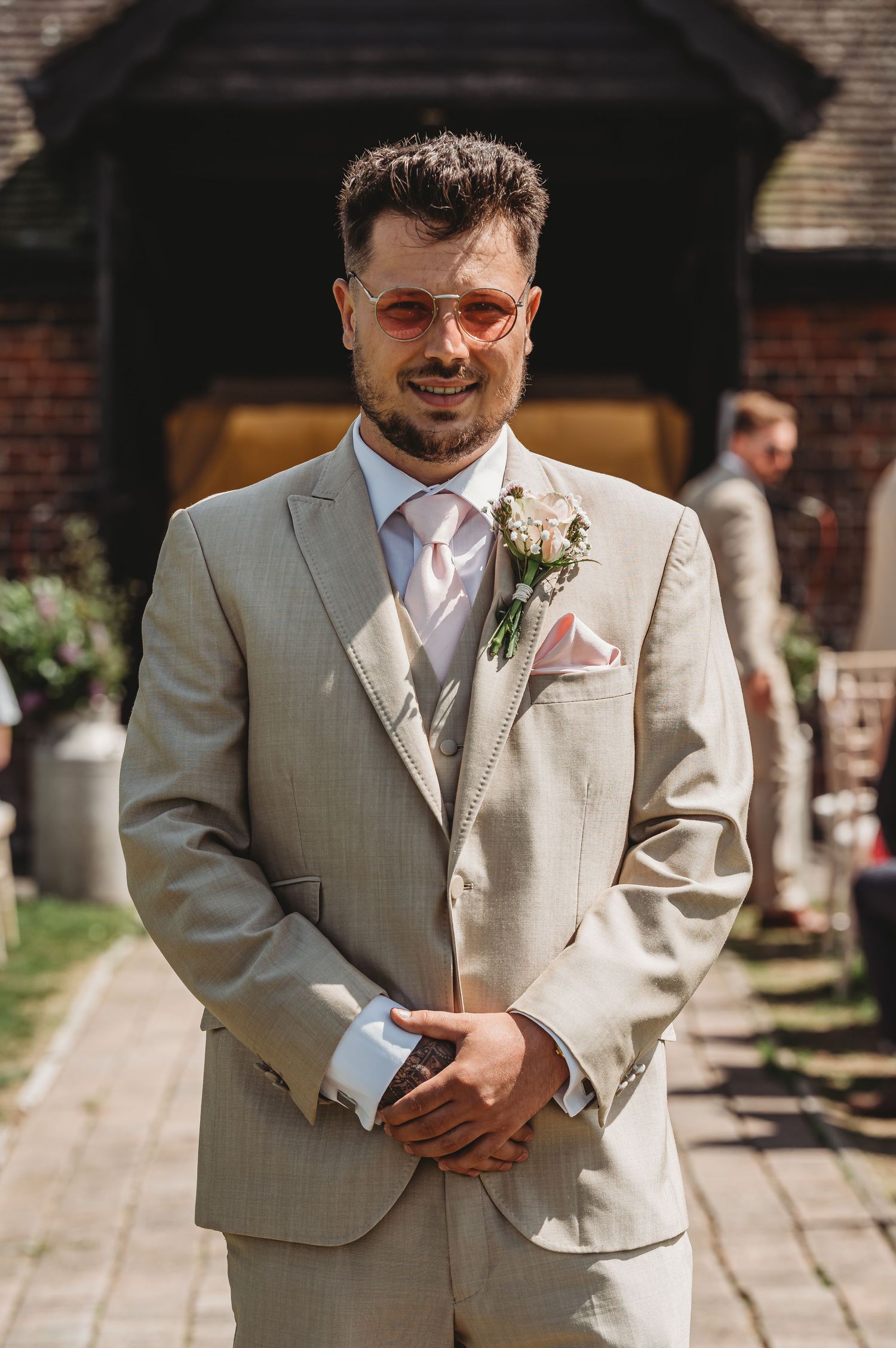 A groom in a light beige suit and glasses smiles while wearing a soft pink tie and floral boutonniere at an outdoor venue.