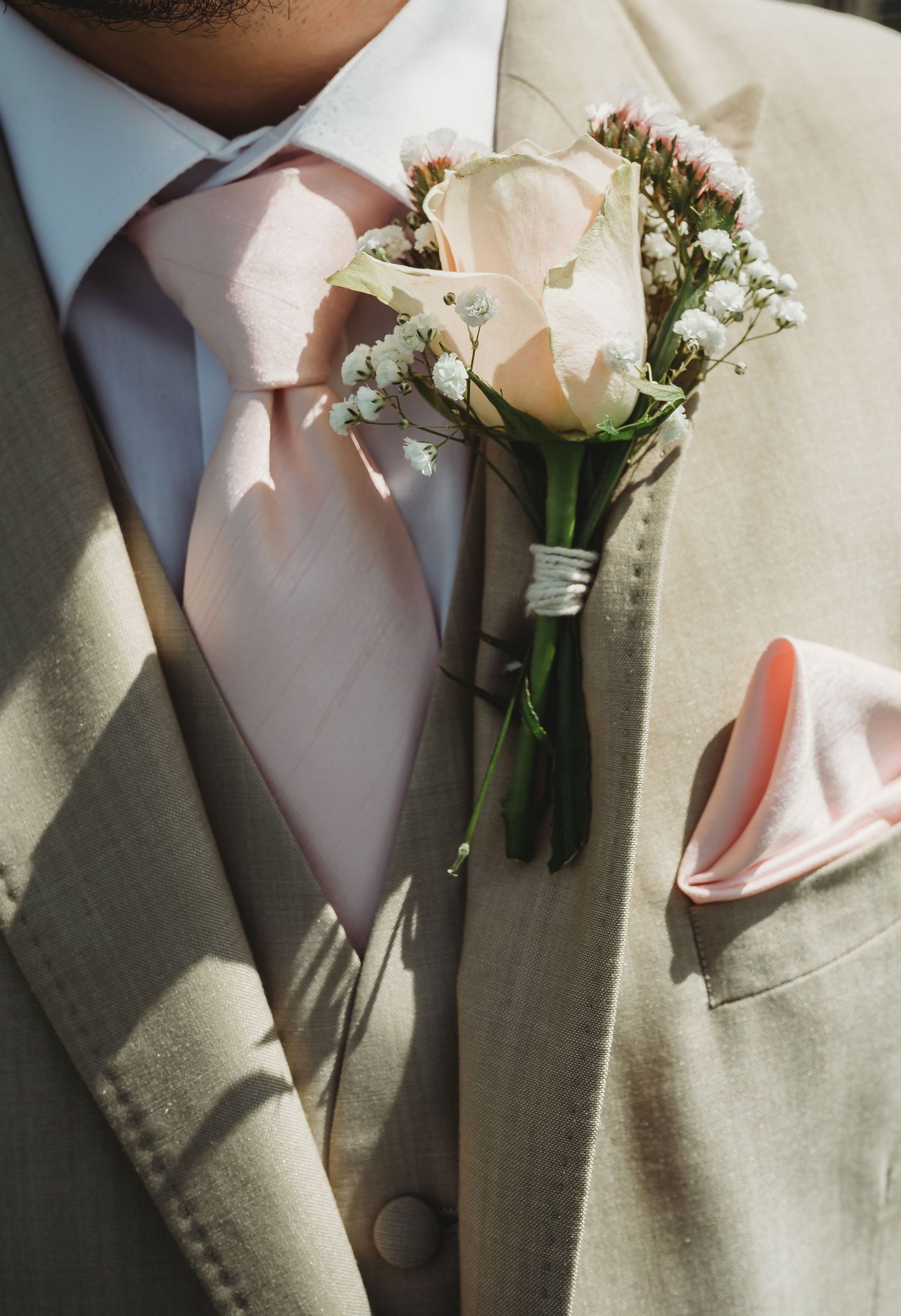 A white rose and baby's breath boutonniere adorns a beige suit jacket with pink tie and matching pocket square.