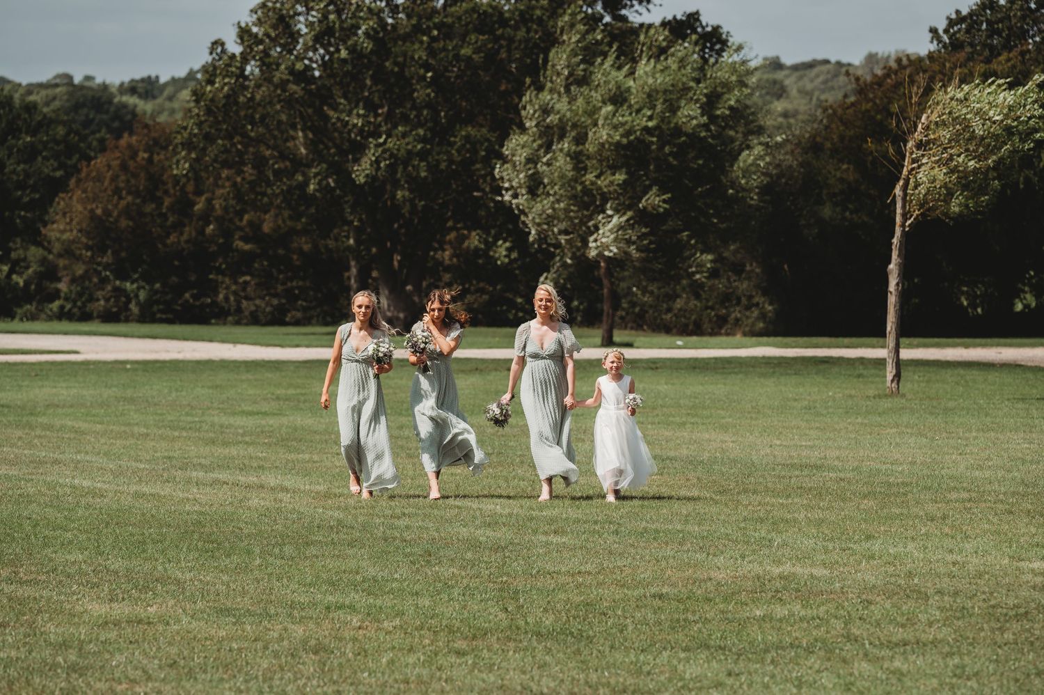 Wedding party wearing white dresses runs joyfully across a sunlit green lawn on a beautiful summer day.