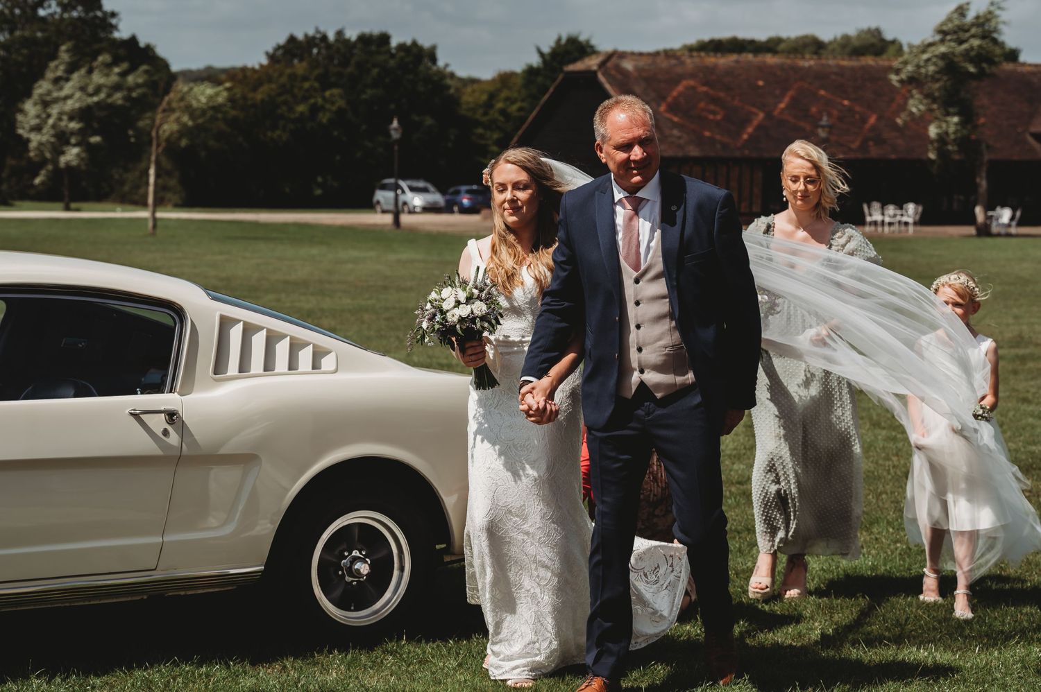Classic white Mustang car parked outside a rustic barn while wedding party members walk in flowing white dresses and suit.