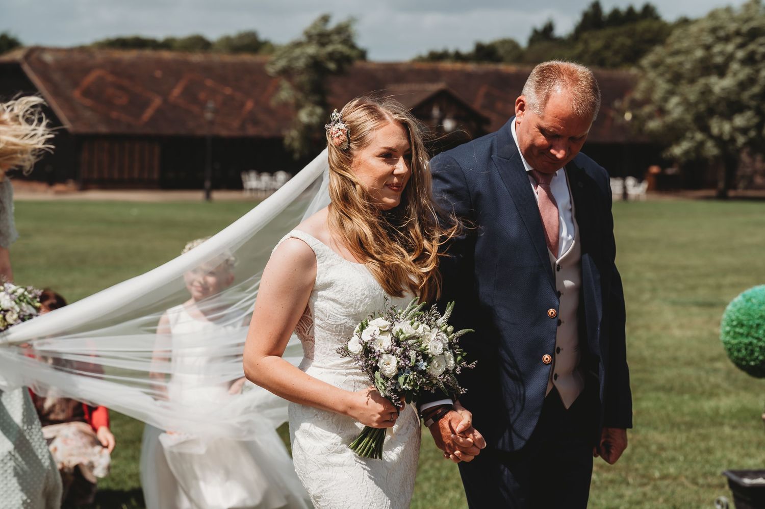 A joyful wedding moment captured outdoors with flowing veil and white floral bouquet against a rustic barn backdrop.