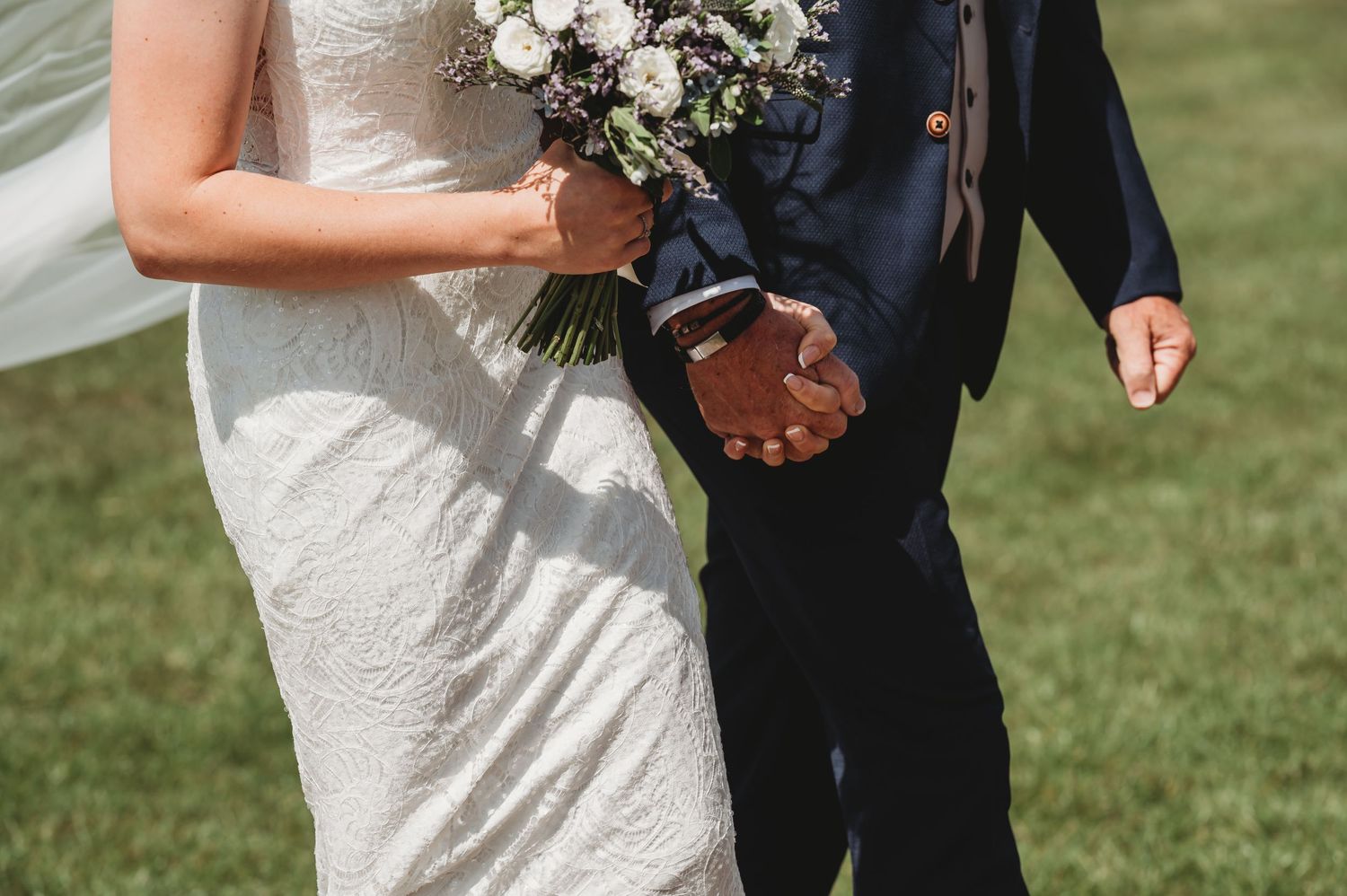 Close-up view of a bride in white dress holding a pink and white bouquet next to someone in navy suit on green lawn.