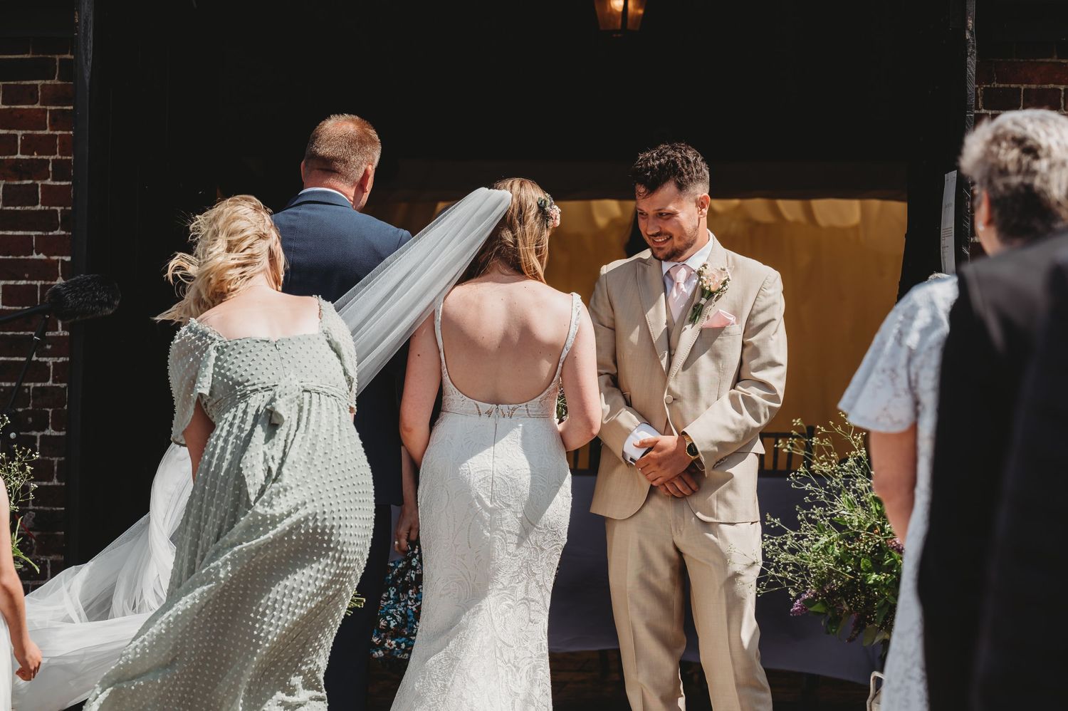 Wedding party members gather outdoors near a brick building as veil flows in the breeze on a sunny day.