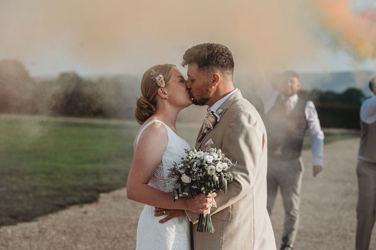 A romantic kiss at sunset between newlyweds in wedding attire with a soft-focus blurred figure in the background.