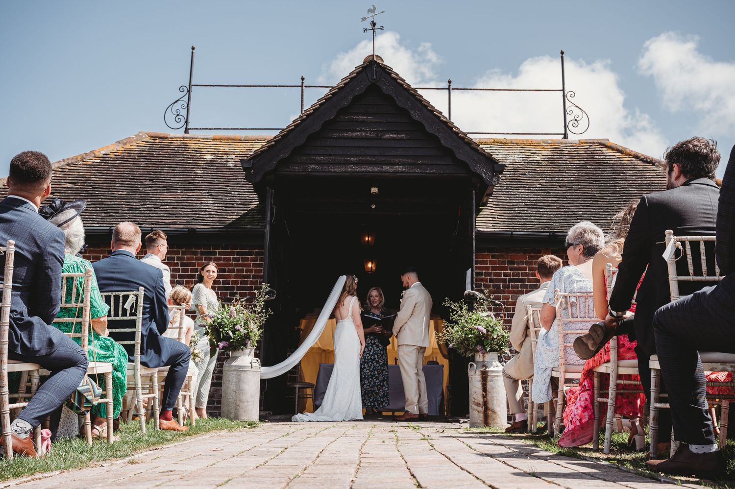 A group of wedding guests watch as two people exit a rustic wooden chapel with a cross on top on a sunny day.