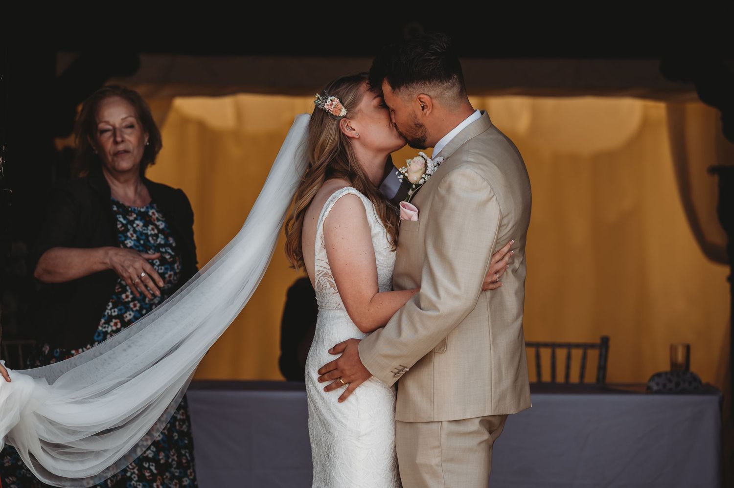 A couple shares their first kiss as newlyweds at their wedding ceremony while a flowing white veil trails behind.
