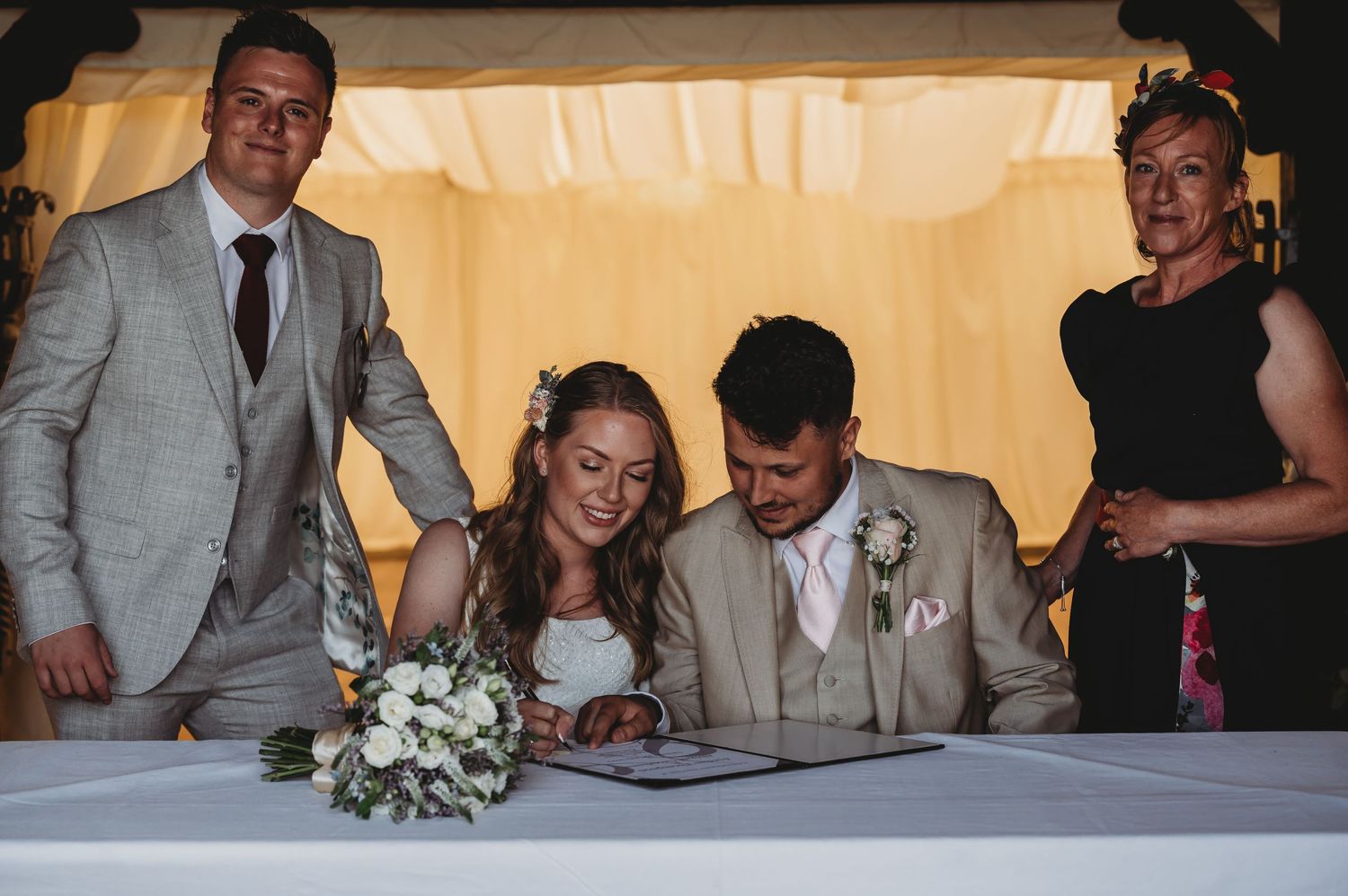 A couple signs their marriage documents at a reception table while guests watch during an indoor wedding ceremony.