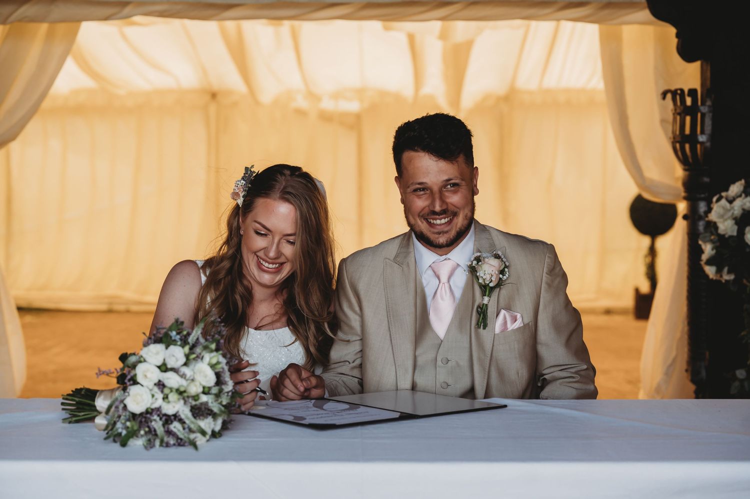 A couple signs documents at a wedding ceremony beneath a light-colored marquee.