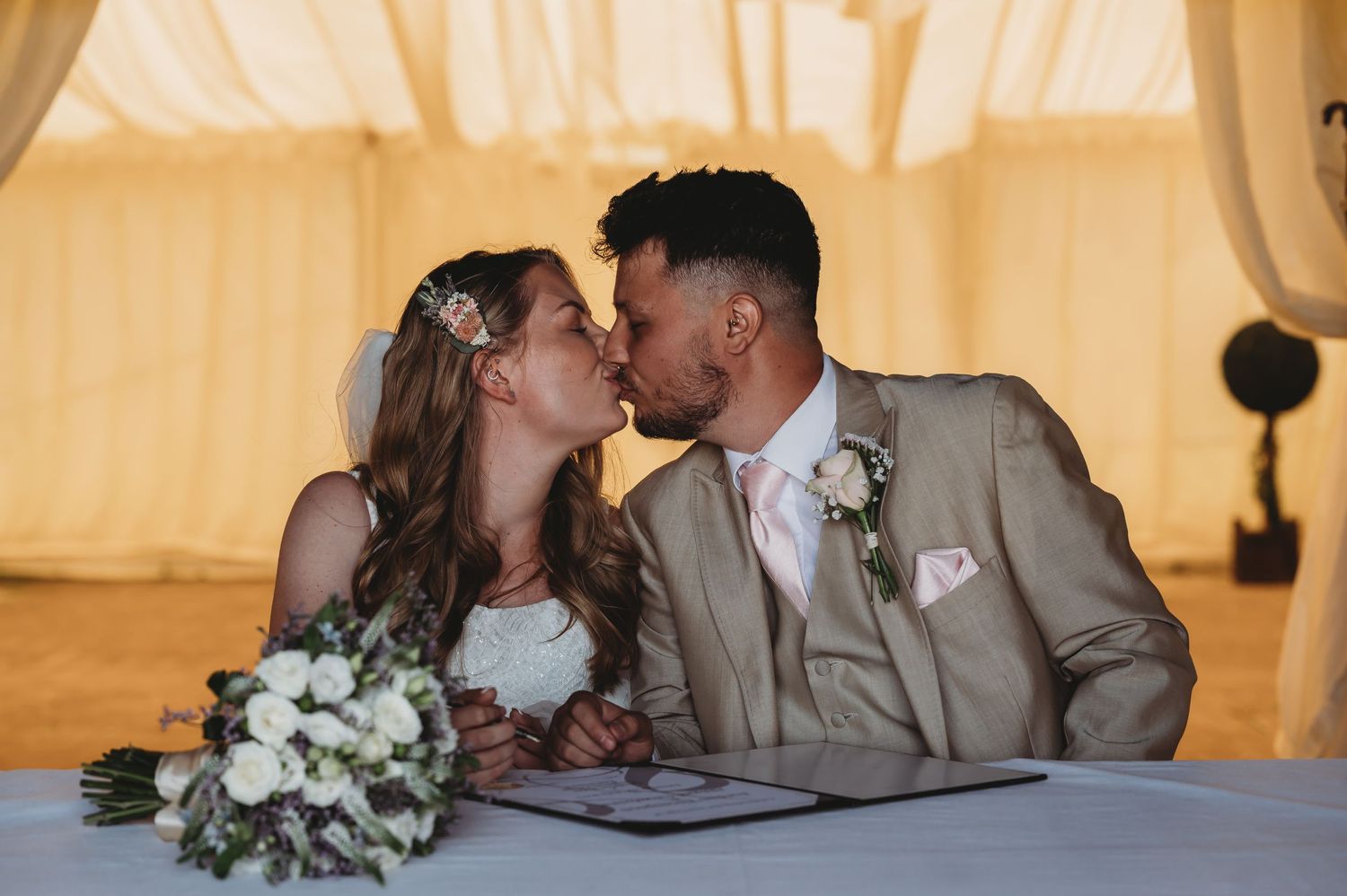 A romantic moment shared between newlyweds at their wedding reception while signing documents with a white bouquet nearby.